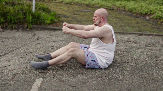 Ein Mann in Unterwäsche sitzt mit einer fahrenden Haltung auf dem Boden. © Screenshot 