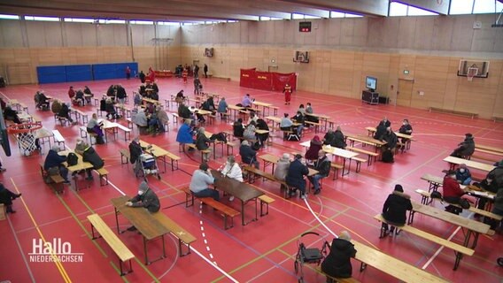 Menschen sitzen in einer Turnhalle an Tischen, Blick von oben. © Screenshot 