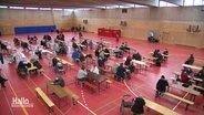 Menschen sitzen in einer Turnhalle an Tischen, Blick von oben. © Screenshot 