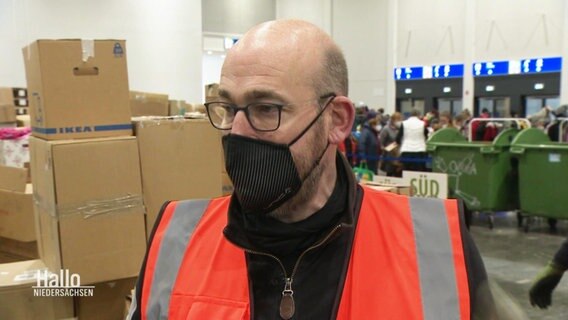 Ein Mann mit Warnweste in einer Halle, im Hintergrund Kartons. © Screenshot 