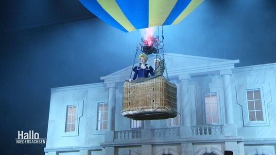 Auf einer Theaterbühne schwebt eine Frau in einem Heißluftballon vor einer klassizistischen Häuserfassade. © Screenshot 