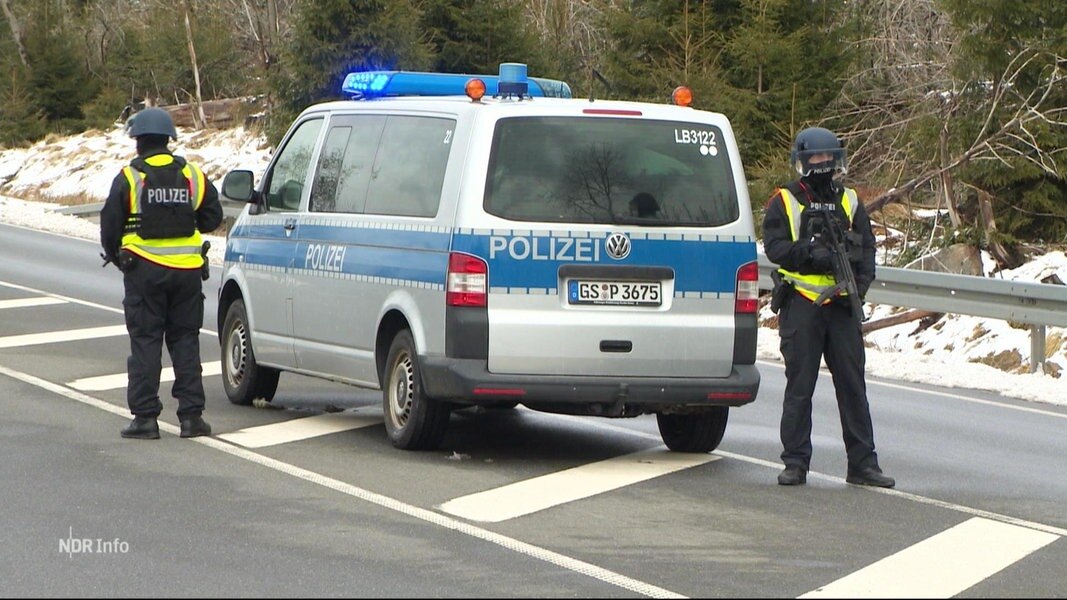 Zwei schwerbewaffnete Polizosten stehen neben ihrem Einsatzfahrzeug auf einer zweispurigen Landstraße.
