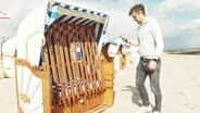 Ein Mann steht mit seinem Smartphone vor einem versperrten Strandkorb © Screenshot 