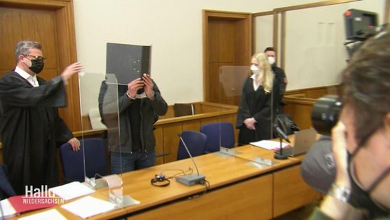 Ein Angeklagter hält sich im Gerichtssaal einen Ordner als Schutz vor sein Gesicht. © Screenshot 