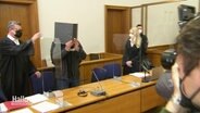 Ein Angeklagter hält sich im Gerichtssaal einen Ordner als Schutz vor sein Gesicht. © Screenshot 