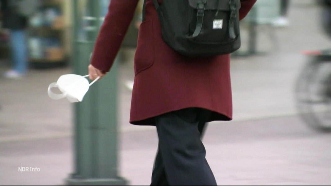 Eine Frau geht über die Straße. In der linken Hand trägt sie lose eine weiße FFP2-Maske.