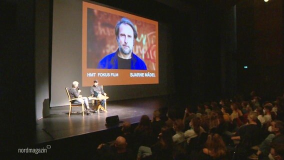 Auf einer Bühne sitzen ein Moderator und der Schauspieler Bjarne Mädel, ein Bild von ihm ist hinter ihnen an die Wand projiziert. © Screenshot 