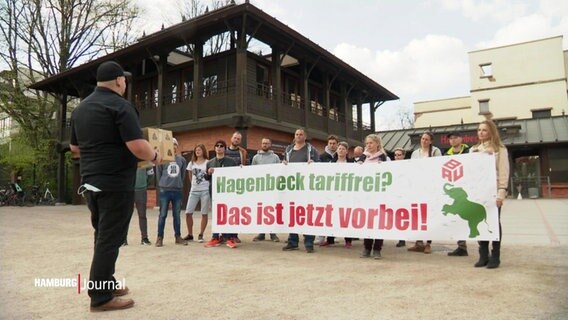 Mitarbeitende von Hagenbecks Tierpark stehen vor dem berühmten Eingangstor und halten ein Banner mit der Aufschrift: "Hagenbeck tariffrei? Das ist jetzt vorbei!" © Screenshot 