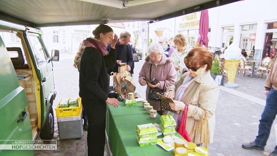 Am Marktstand stehen einige Interessierte und kaufen ein. © Screenshot 