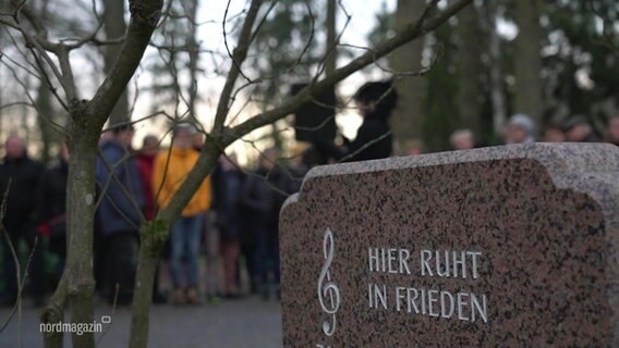 Eine Gruppe von Menschen auf einem Friedhof, im Vordergrund ein Grabstein mit der Aufschrift: "Hier ruht in Frieden" © Screenshot 