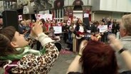 Viele Menschen stehen auf dem Beatles Platz auf der Hamburger Reeperbahn. Einige halten Schilder mit Herzen empor. In der Mitte des Bildes, singt Leon Mancilla, der Sänger der Band "Salamanda", in ein Mikrofon. © Screenshot 