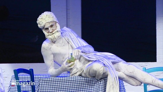 Ein Schauspieler, verkleidet als Zeus-Statute, liegt halb auf einem Restauranttisch. © Screenshot 