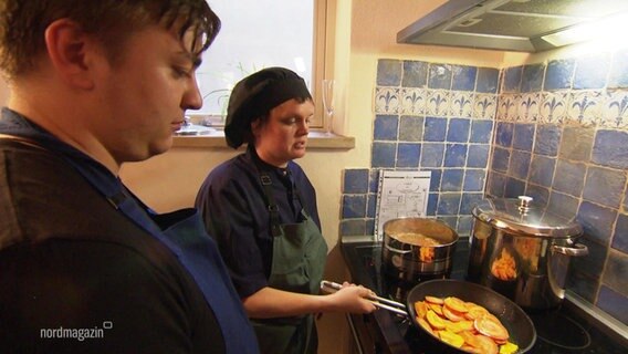 Jule Klinger zeigt einem Teilnehmer ihres veganen Kochkurses in Rostock ein Gericht in einer Pfanne. © Screenshot 