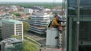 Von einem hängenden Korb aus, putzen zwei Männer Fenster. © Screenshot 
