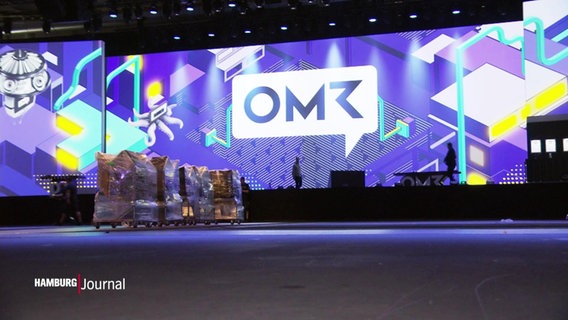 Noch wird aufgebaut: Eine große Bühne mit Leinwand und dem Logo von OMR, den Online Marketing Rockstars, in den Messehallen. © Screenshot 