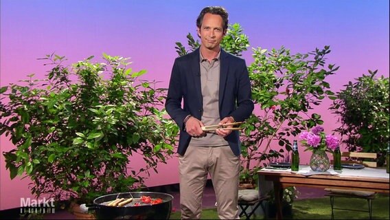 Moderator Jo Hiller steht in einem Set mit Pflanzen, einem Grill und einem gedeckten Gartentisch, die Grillzange in der Hand. © Screenshot 