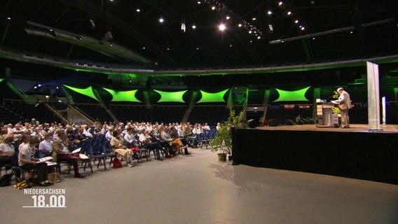 In einer größeren Kongresshalle sitzen mehrere Menschen und hören einer Rede zu. © Screenshot 
