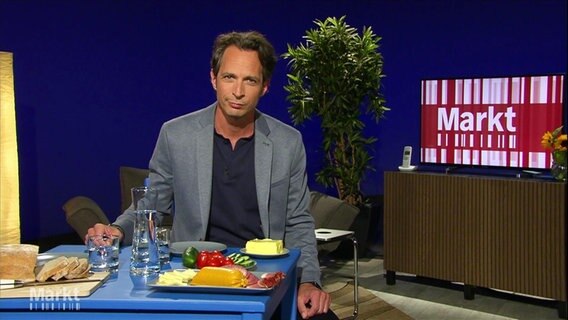Moderator Jo Hiller sitzt an einem gedeckten Tisch mit Lebensmitteln für ein typisches Abendbrot. © Screenshot 