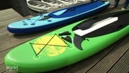 Ein grünes und ein blaues SUP, jeweils mit Pinnen und noch nicht montierten Finnen, liegen auf einem Steg. © Screenshot 