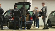 Polizisten und weitere Personen stehen an einem dunklen Kombi, ein Mann lädt einen großen Karton in den Kofferraum. © Screenshot 