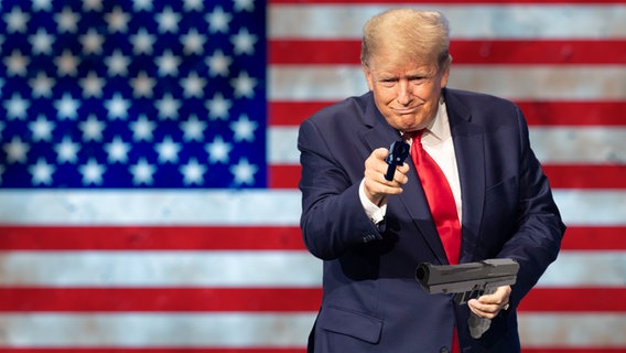 Trump mit Waffe, im Hintergrund die US-Fahne.  