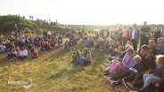 Eine Gruppe Menschen sitzt in den Dünen und singt. © Screenshot 