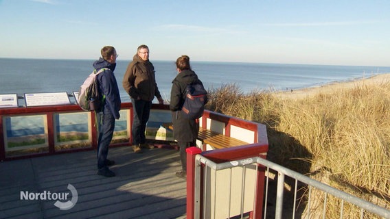 Drei Personen stehen auf einer Aussichtsplattform und schauen auf den Strand. © Screenshot 