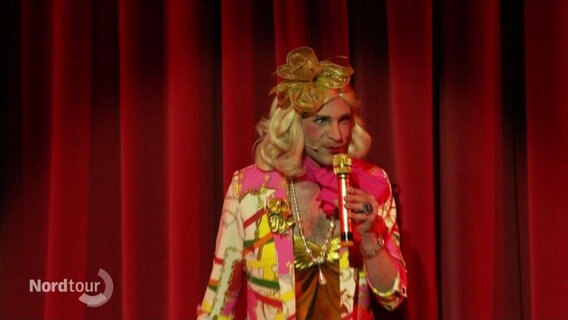 Ein Mann in goldenem Kleid steht auf einer Bühne vor rotem Vorhang. © Screenshot 