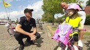 Ein Polizist befragt ein kleines Kind auf einem Fahrrad. © Screenshot 