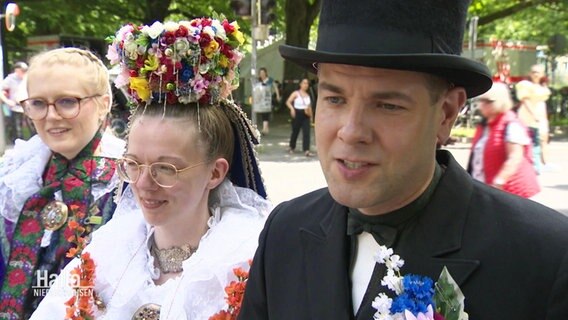 Carina Bahlburg (mitte) in einem traditionellen Brautgewand, mit Blumenkrone. Rechts von ihr steht Carsten Bahlburg, in der Mode eines Bräutigams. © Screenshot 