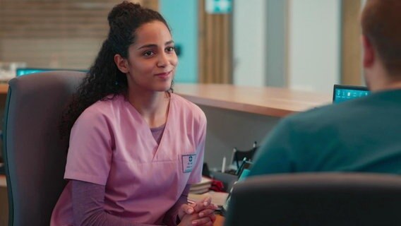 Eine junge Frau in violetter Krankenhaus-Arbeitskleidung lächelt jemandem zu, der nur im Anschnitt zu sehen ist. © Screenshot 
