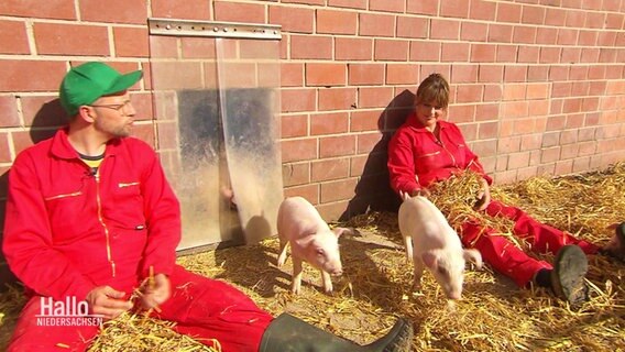 Reporter Andi Gervelmeyer und Schweinehalterin Gabi Mörixmann sitzen vor einem Stall auf dem Boden. © Screenshot 