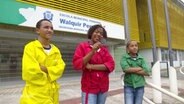 Drei Kinder aus Brasilien stehen nebeneinander und schauen in die Kamera. Das Kind in der Mitte hält eine Lupe in der Hand. © Screenshot 
