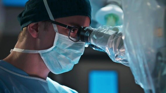 Ein Arzt mit Mundschutz im OP: Er schaut durch ein Mikroskop. © Screenshot 