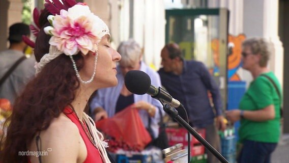 Eine Frau mit Blumenschmuck auf dem Kopf, singt in ein Mikrophon. © Screenshot 