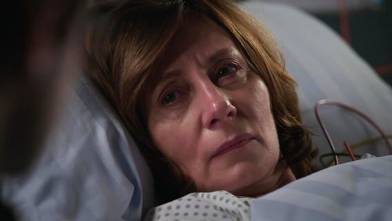 Eine Frau liegt in einem Krankenhausbett, Großaufnahme ihres Gesichts. Sie schaut jemanden außerhalb des Bildes an; ihr Blick ist erschöpft und traurig. © Screenshot 