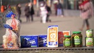 In einer Fußgängerzone stehen mehrere Lebensmittel, wie Toast, Taschentücher und Zwieback, im Fokus. © Screenshot 