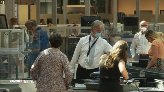 Szene an der Sicherheitskontrolle am Flughafen: Passagiere legen Gepäck auf das Förderband, ein Mitarbeiter der Sicherheitskontrolle mit Mundschutz. © Screenshot 