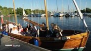 Mehrere Menschen sitzen in der Abendsonne auf einem Holzsegelboot an der Außenalster. © Screenshot 