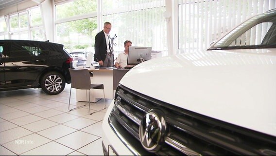Blick auf die Motorhaube eines weißen VWs. Im Hintergrund sprechen zwei Männer in Anzügen miteinander in einem Autohaus. © Screenshot 