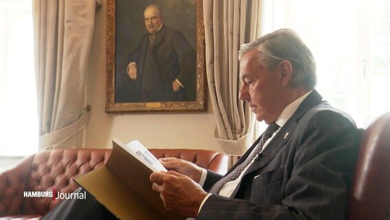 Der Präsident des Hamburger Übersee-Clubs, Michael Behrendt, sitzt in einem Ledersessel und blättert in einem Buch. © Screenshot 