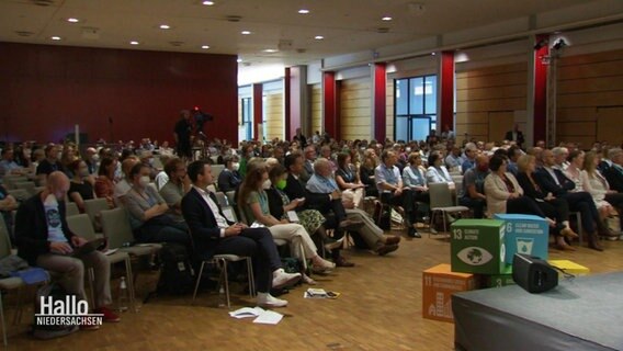 In einem Konferenzsaal sitzen viele Menschen in mehreren Stuhlreihen. © Screenshot 
