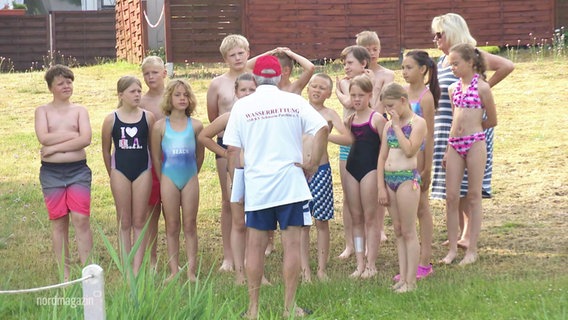 Ein älterer Mann in Bademeister-Klamotten steht vor einer Gruppe von Kindern in Schwimmkleidung. © Screenshot 