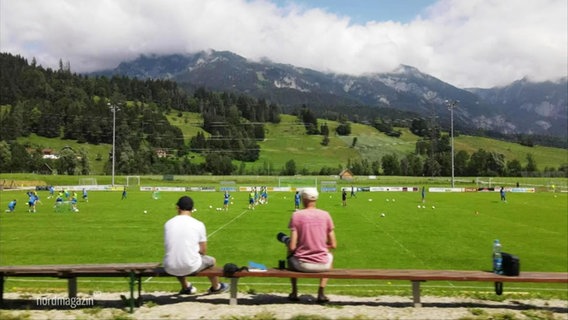 Zwei Personen sitzen auf einer Bank am Rand eines reisigen in den Bergen gelegenen Fußballfeldes. © Screenshot 