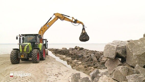 Ein Bagger hebt einen schweren Stein um ihn auf einer Steinpallisade an einem Strand abzusetzen. © Screenshot 