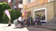 Eine Familie sitzt beim Eisessen vor einem Café, die Mutter im Rollstuhl. © Screenshot 