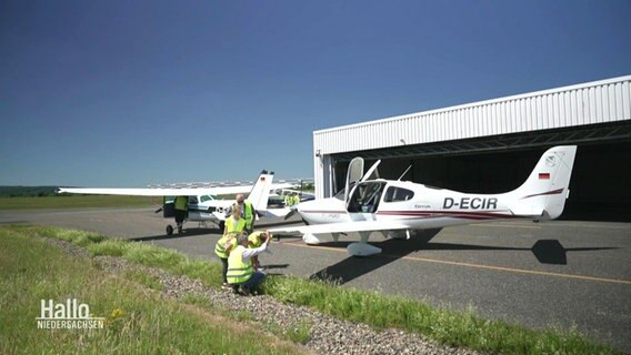 Eine Lehrer steht mit Schülerinnen in Warnweste vor einem kleinen Segelflugzeug. © Screenshot 
