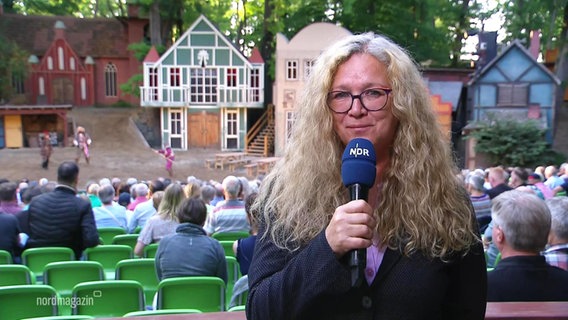 Reporterin Heike Becker berichtet live vor Ort, im Hintergrund ist die Bühne zu sehen. © Screenshot 