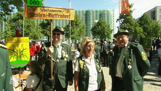 Beim Schützenausmarsch in Hannover stehen 3 Personen in traditioneller Tracht und halten ein Schild ihres Schützenvereins hoch. © Screenshot 