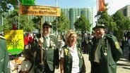 Beim Schützenausmarsch in Hannover stehen 3 Personen in traditioneller Tracht und halten ein Schild ihres Schützenvereins hoch. © Screenshot 